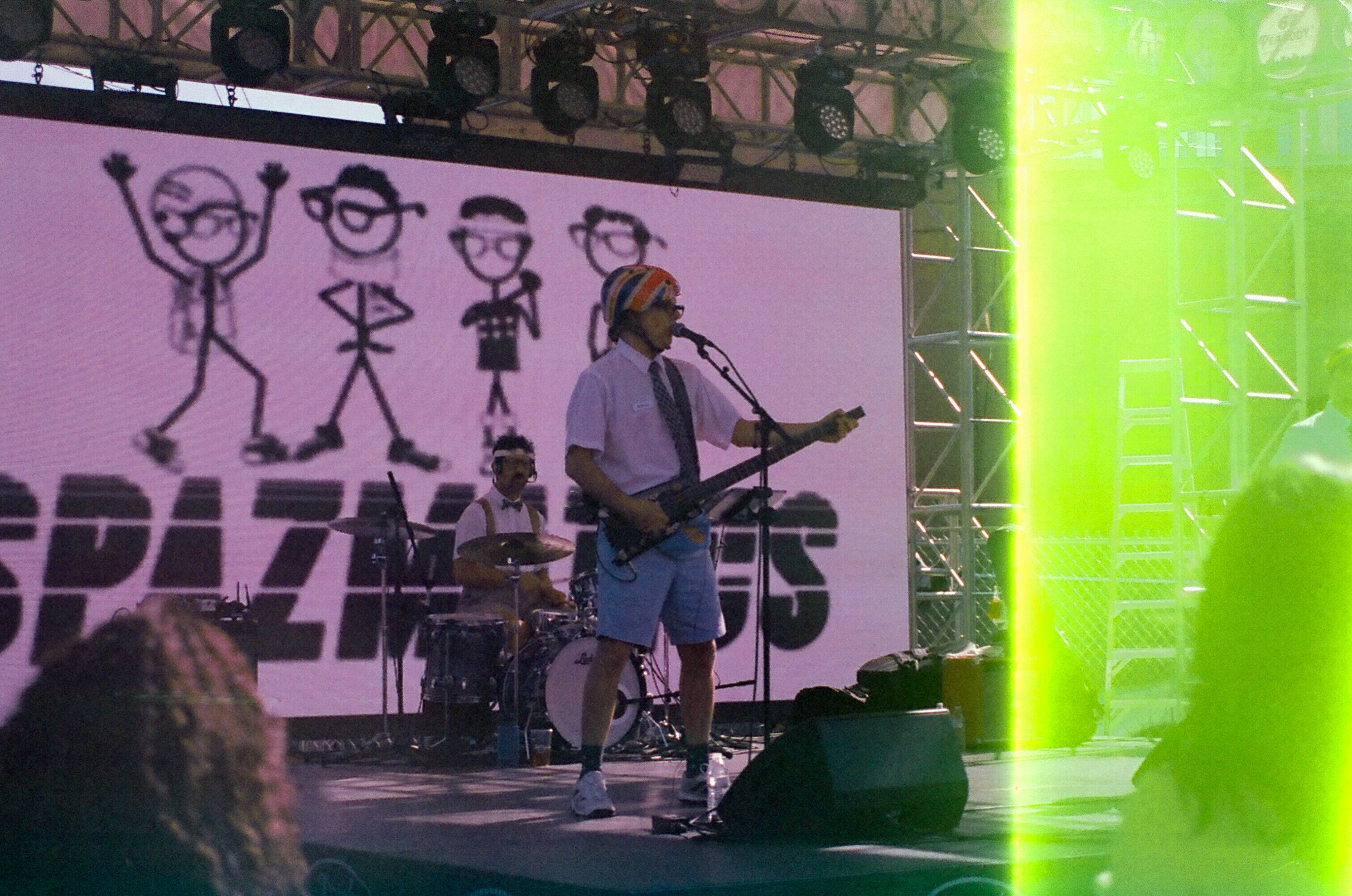 Photo of a band playing on stage. A singer with an electric guitar is singing into a microphone stand, and wearing a white button-down shirt and blue shorts. A drummer is behind him with a similar shirt, suspenders, and a bowtie. The stage backdrop shows a drawing of four stick figures with nerdy-looking clothes and glasses, and the name "SPAZMATICS." There is a strong green vertical light leak down the right quarter of the photo.
