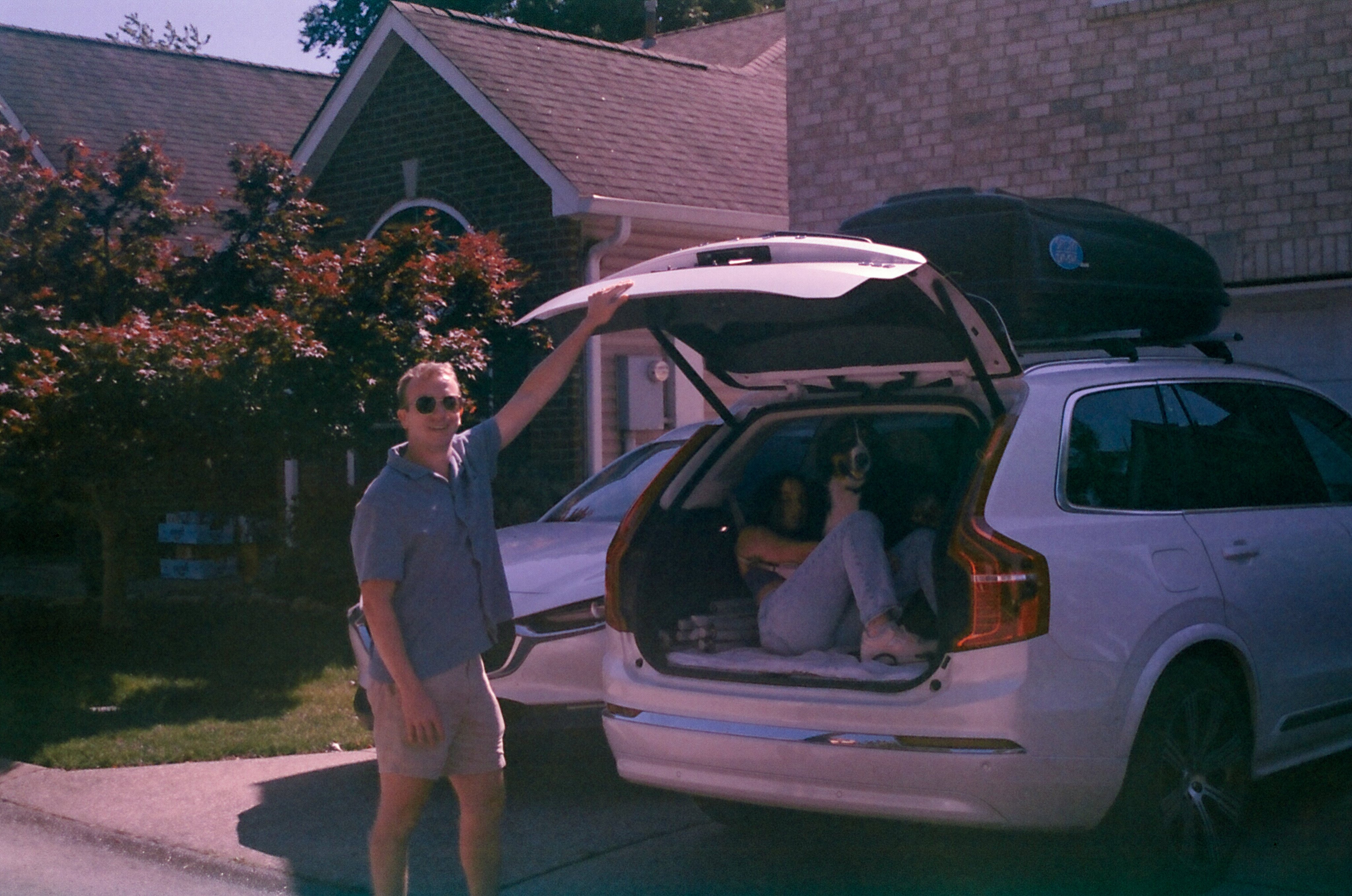 Photo of a white SUV sitting in a driveway with the trunk open. A man wearing a short-sleeve button down shirt, shorts, and sunglasses is standing behind the car with his left hand holding the open trunk. A woman is sitting in the trunk with a Bernese Mountain Dog.