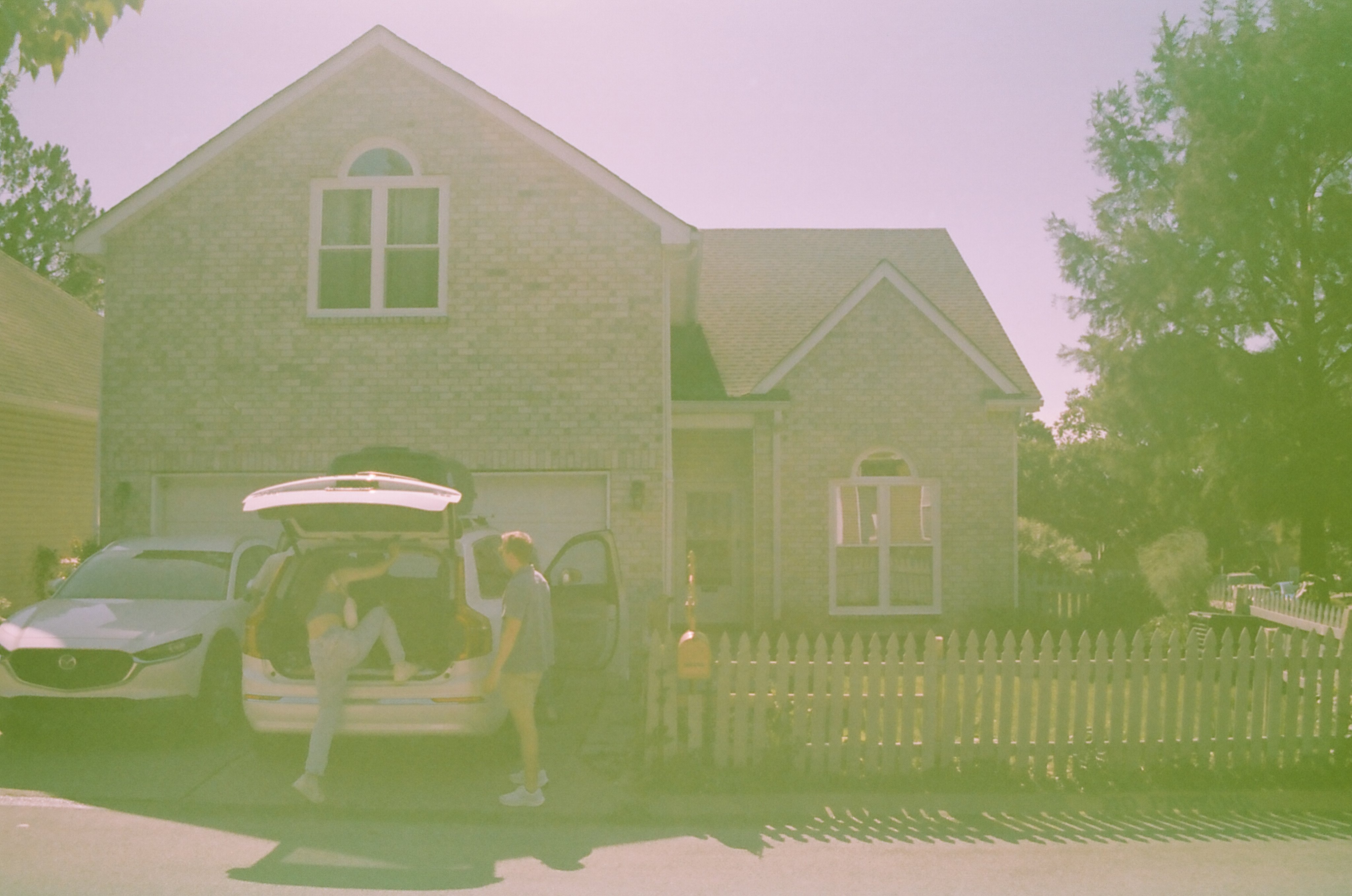 Photo of two people behind an SUV with its trunk open, which is parked in the driveway of a small two-story house with a gray-brick facade. The person on the left has one leg up on the bumper as they climb into the trunk, and the person on the right is standing upright and watching the first. The entire photo has a strong green color cast.