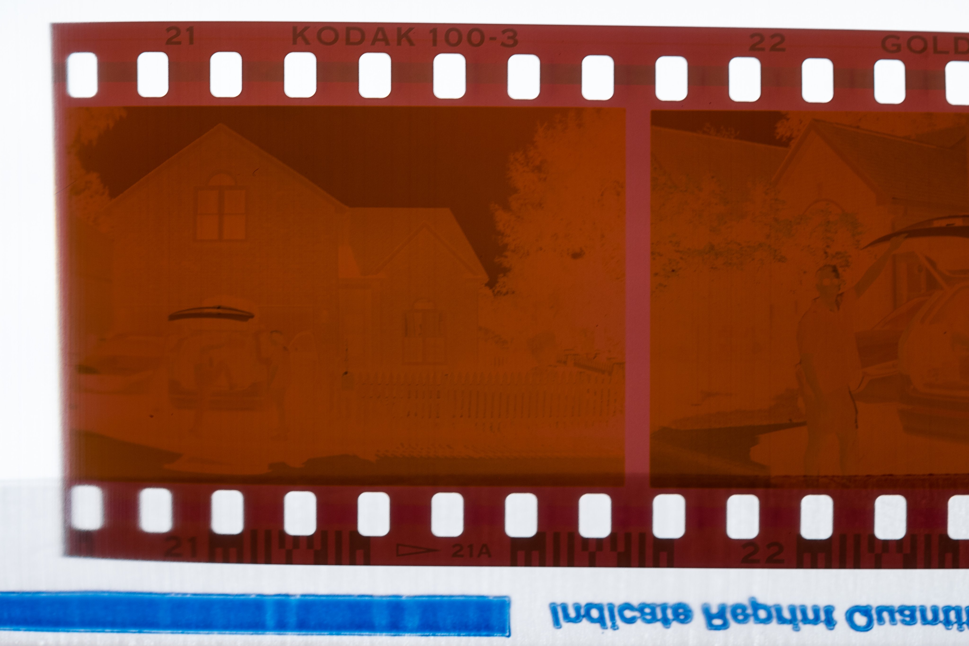 Close-up of a film negative, showing little detail, though a house and cars in the driveway with two people can be made out. The film border reads "21 KODAK 100-3 22 GOLD"
